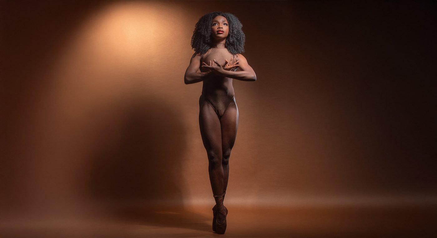 Dancer en pointe in a nude bodysuit under a warm spotlight