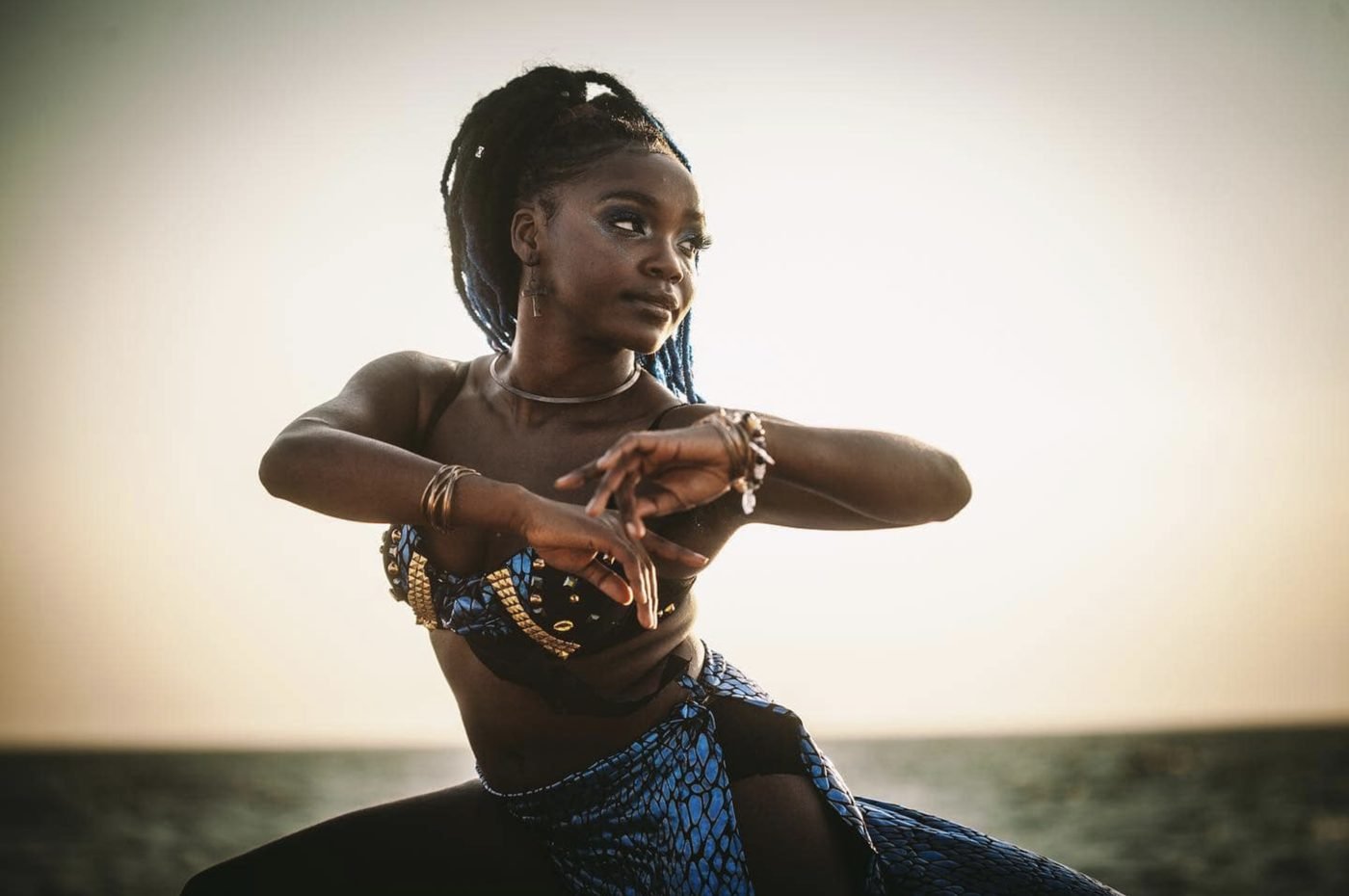 Dancer in blue African print with long braids posing by the sea at sunset