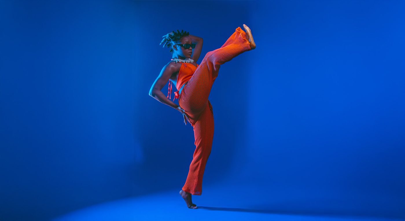 Dancer kicking in an orange mesh outfit against a blue backdrop