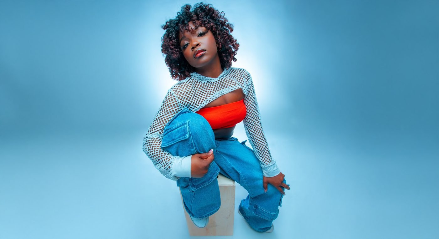 Dancer in a mesh top and blue jeans crouching against a blue backdrop